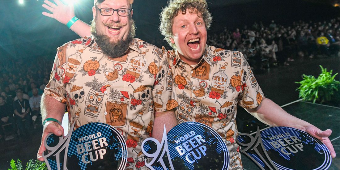 Two men in matching shirts pose with three World Beer Cup awards