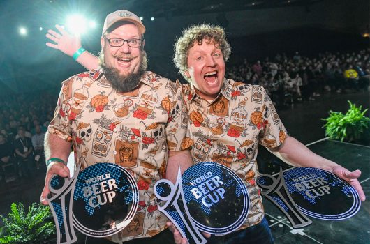 Two men in matching shirts pose with three World Beer Cup awards